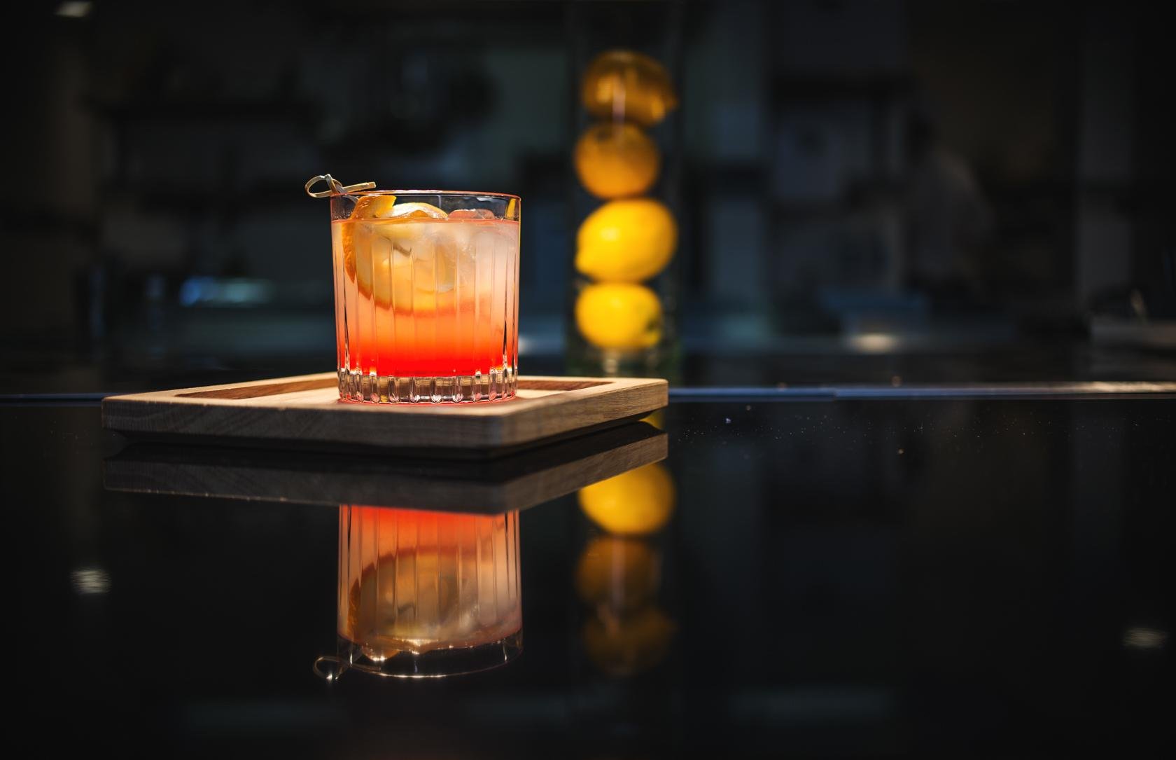 Old fashioned cocktail on the bar, on a wooden tray with lemons in the background. The Old Fashioned is a cocktail made by muddling sugar with bitters, then adding alcohol, such as whiskey or brandy, and a twist of citrus rind